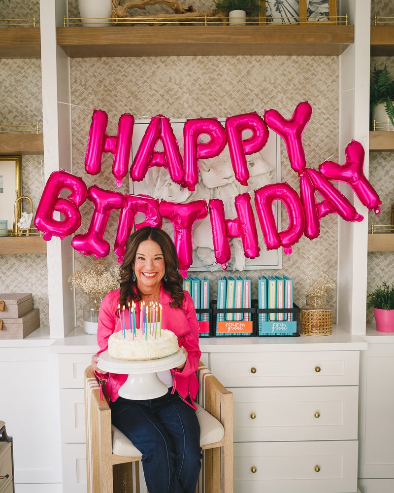 Jamie holding a candle-lit birthday cake in front of a hot-pink HAPPY BIRTHDAY balloon banner